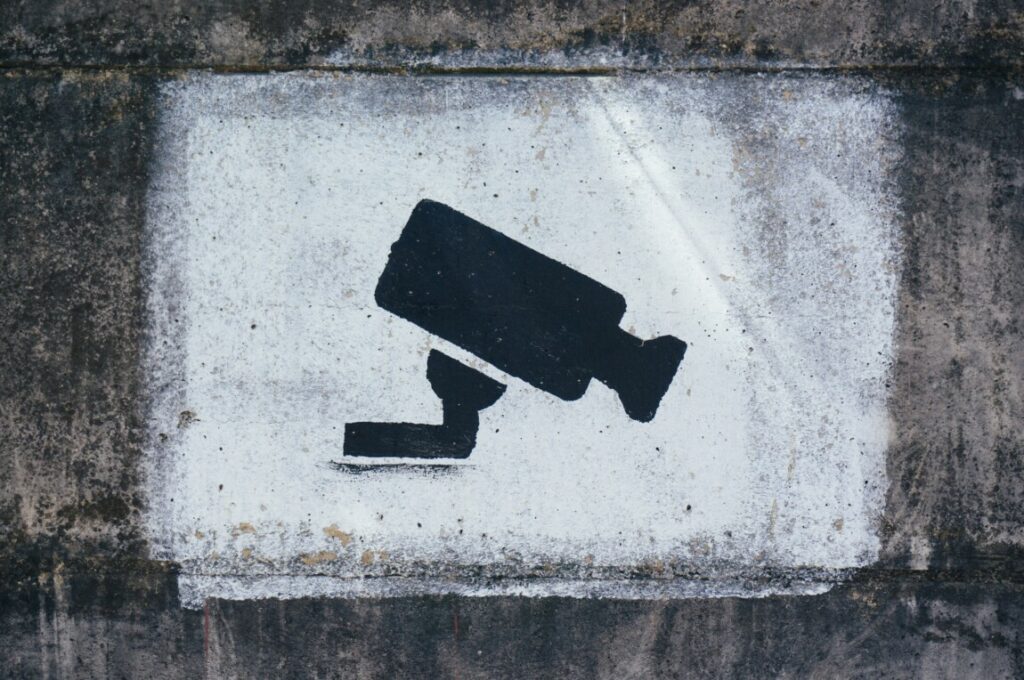 The outline of a CCTV camera, in black, surrounded by white paint on a concrete wall